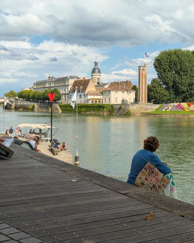 A riverside promenade with wooden decking and stone paving, featuring people relaxing on benches and the riverbank, with a view of a bridge, historic buildings including a church with a dome, and a French flag flying in the background.