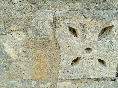 A close-up photograph of a weathered stone block featuring a carved cross with four petal-shaped openings, set within a rough stone wall.