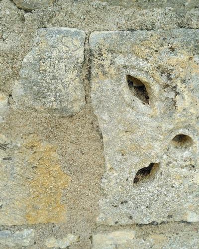 A close-up photograph of a weathered stone block featuring a carved cross with four petal-shaped openings, set within a rough stone wall.