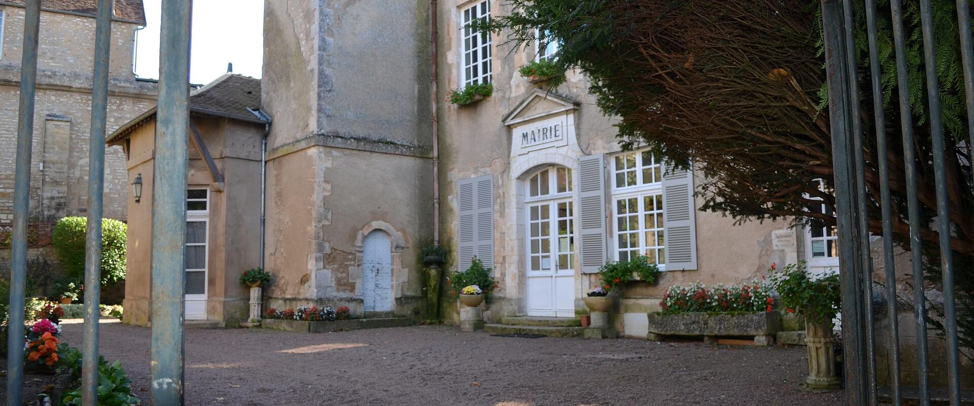 The image shows the exterior of a stone building with a sign above the entrance reading "MAIRIE" (French for town hall), featuring shuttered windows, flower boxes, and a gravel courtyard viewed through metal gates.
