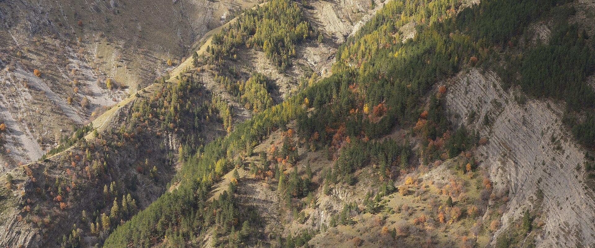1. A mountainous landscape in the French Alps showing steep, rocky slopes with patches of coniferous and deciduous trees displaying autumn colors, a small white building nestled in a valley, and cloud-covered peaks in the background.