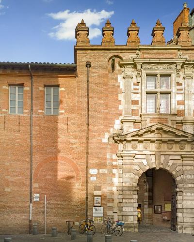 The image shows the main entrance facade of a historic brick building with a prominent stone archway, decorative Renaissance-style stonework around the doorway and upper window, and a crenellated parapet; a street sign reading "PLACE D'ASSEZAT" is