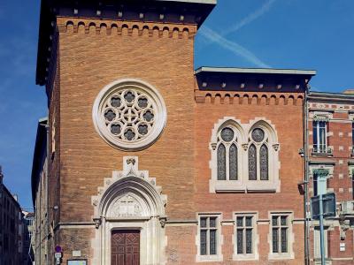 A red-brick building with Gothic Revival architectural features, including a large rose window, pointed-arch windows, and an ornate stone entrance, situated on a city street under a blue sky.