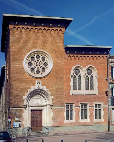 A red-brick building with Gothic Revival architectural features, including a large rose window, pointed-arch windows, and an ornate stone entrance, situated on a city street under a blue sky.