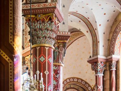 The image shows the interior of a Romanesque church featuring brightly painted columns with sculpted capitals, arched ceilings decorated with stars and birds, and a chandelier.