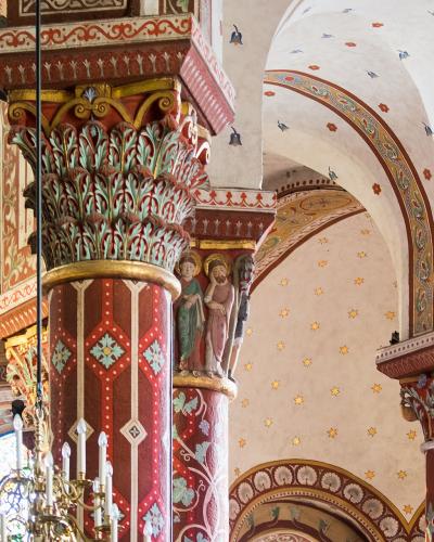 The image shows the interior of a Romanesque church featuring brightly painted columns with sculpted capitals, arched ceilings decorated with stars and birds, and a chandelier.