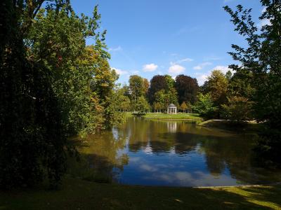 A scenic view of a park featuring a calm pond reflecting the blue sky and surrounding trees, with a white classical-style pavilion visible on the far bank.