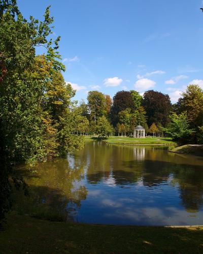 A scenic view of a park featuring a calm pond reflecting the blue sky and surrounding trees, with a white classical-style pavilion visible on the far bank.