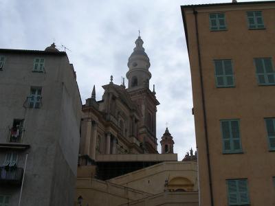 The image shows the Basilique Saint-Michel in Menton, France, identifiable by its distinctive pinkish facade, prominent bell tower, and the surrounding steep, terraced urban architecture typical of the town.