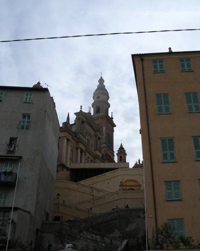 The image shows the Basilique Saint-Michel in Menton, France, identifiable by its distinctive pinkish facade, prominent bell tower, and the surrounding steep, terraced urban architecture typical of the town.
