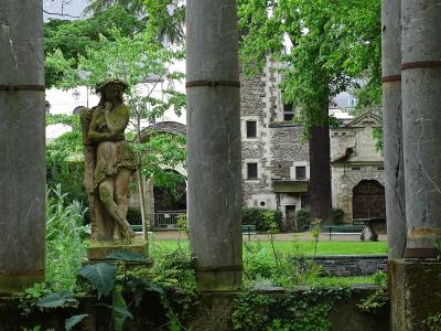 A view of a historic stone courtyard featuring a moss-covered statue of a woman, large concrete pillars in the foreground, and medieval-style stone buildings with arched doorways and windows in the background.