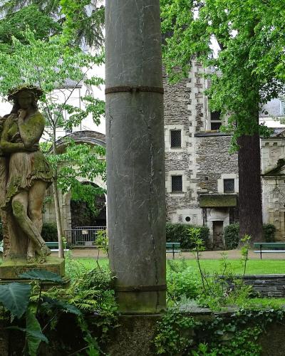 A view of a historic stone courtyard featuring a moss-covered statue of a woman, large concrete pillars in the foreground, and medieval-style stone buildings with arched doorways and windows in the background.