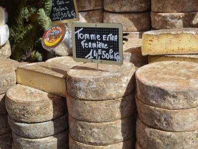 A close-up of stacked wheels of artisanal cheese at a market stall, featuring a handwritten sign in French for "Tomme Extra Fermière" priced at 14,50€/kg and another sign for "des Moines" at 22€/kg.