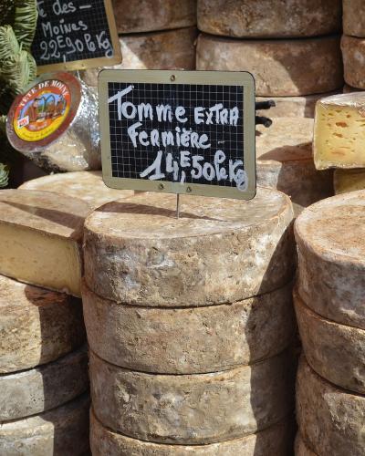 A close-up of stacked wheels of artisanal cheese at a market stall, featuring a handwritten sign in French for "Tomme Extra Fermière" priced at 14,50€/kg and another sign for "des Moines" at 22€/kg.