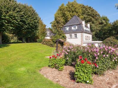 A sunlit garden scene featuring a manicured lawn, a flower bed with pink and red dahlias in the foreground, and a traditional white house with a slate roof and dormer windows nestled among mature trees in the background.