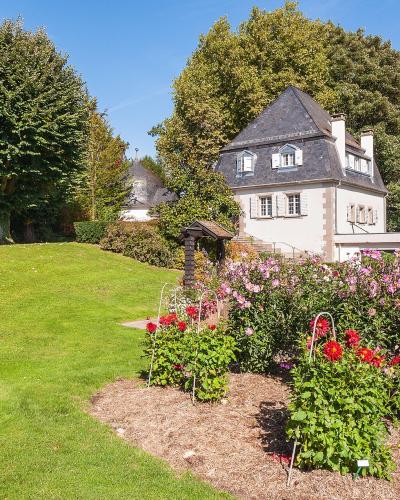 A sunlit garden scene featuring a manicured lawn, a flower bed with pink and red dahlias in the foreground, and a traditional white house with a slate roof and dormer windows nestled among mature trees in the background.