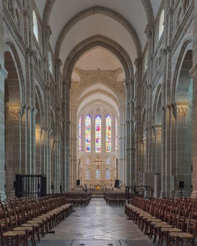 The interior nave of a large Romanesque cathedral, showing rows of wooden chairs facing an altar with stained glass windows under stone arches and columns.