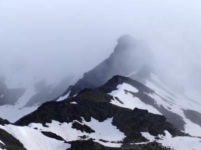 A foggy, high-altitude mountain peak with patches of snow on dark rocky slopes, partially obscured by low clouds or mist.