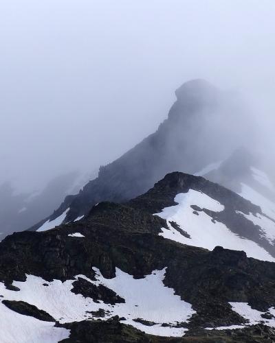 A foggy, high-altitude mountain peak with patches of snow on dark rocky slopes, partially obscured by low clouds or mist.