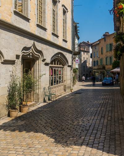 A sunlit, narrow cobblestone street in a historic French village, flanked by stone buildings with arched windows and shopfronts; visible signage includes "VEIL" on the left and a distant pink sign reading "Halle" on the right, with pedestrians and