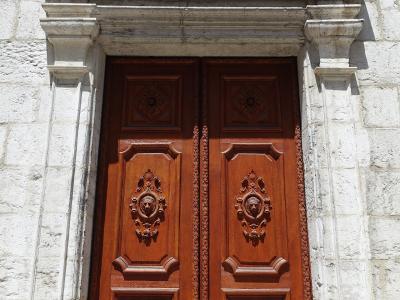 A close-up photograph of a pair of ornate, carved wooden double doors set within a white stone archway featuring a triangular pediment and fluted pilasters.