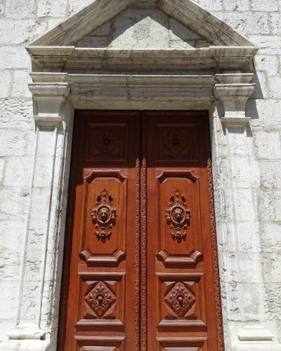 A close-up photograph of a pair of ornate, carved wooden double doors set within a white stone archway featuring a triangular pediment and fluted pilasters.