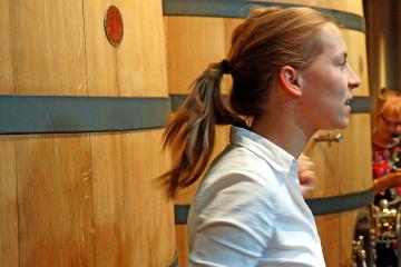 1. A woman in a white shirt stands in profile next to large wooden wine fermentation tanks inside a winery, with another person visible in the background.