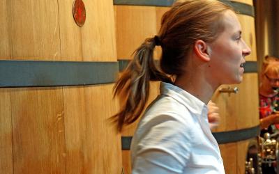 A woman in a white shirt stands in profile next to large wooden wine fermentation tanks inside a winery, with another person visible in the background.