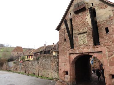 A stone gatehouse with a large arched entryway, part of a fortified wall, with a coat of arms carved above the arch and a view of traditional half-timbered houses and vineyards in the background.