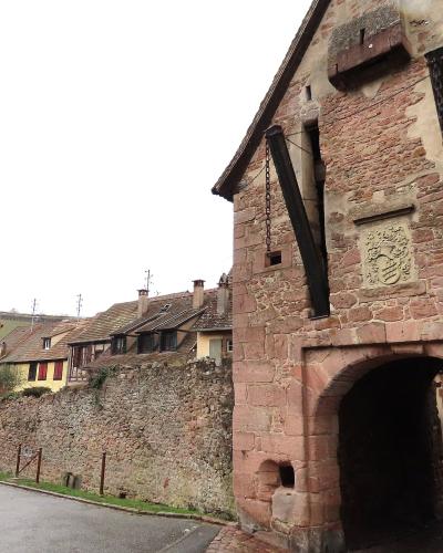 A stone gatehouse with a large arched entryway, part of a fortified wall, with a coat of arms carved above the arch and a view of traditional half-timbered houses and vineyards in the background.