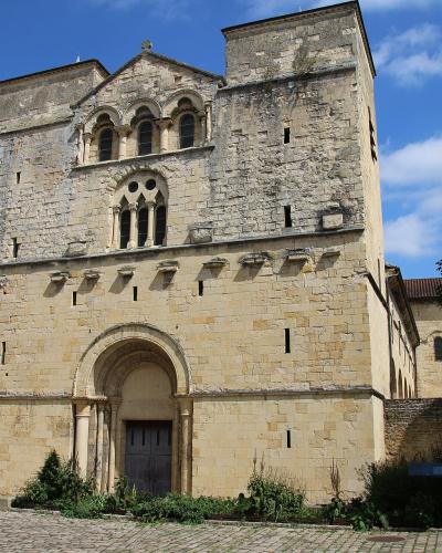 A low-angle photograph of a large, light-colored stone church facade featuring a central arched doorway, a rose window, and three smaller arched windows above, set against a blue sky with a residential building visible to the left.