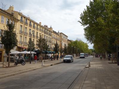 A wide, tree-lined urban boulevard with yellow stucco buildings featuring shuttered windows and wrought-iron balconies on the left, outdoor café seating under umbrellas, pedestrians, parked vehicles including a police van, and a paved pedestrian w