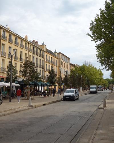 A wide, tree-lined urban boulevard with yellow stucco buildings featuring shuttered windows and wrought-iron balconies on the left, outdoor café seating under umbrellas, pedestrians, parked vehicles including a police van, and a paved pedestrian w
