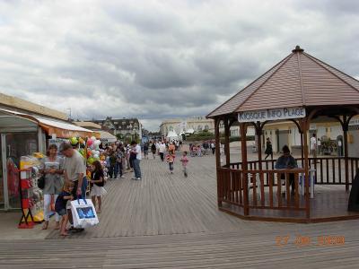 A wooden boardwalk promenade with people walking, a gazebo labeled "KIOSQUE INFO PLAGE," and beachside shops under an overcast sky.