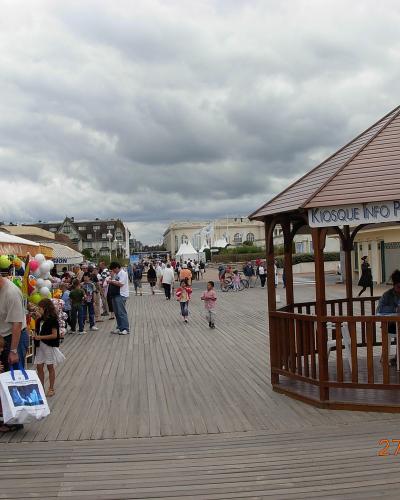 A wooden boardwalk promenade with people walking, a gazebo labeled "KIOSQUE INFO PLAGE," and beachside shops under an overcast sky.