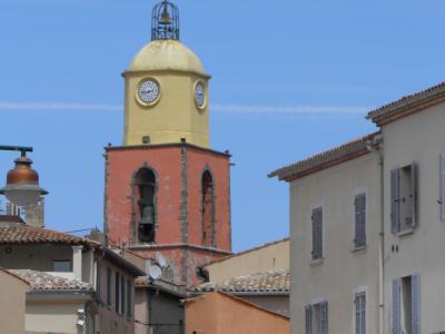 A tall, distinctive bell tower with a salmon-pink lower section and a yellow domed top featuring clocks and an open belfry, rising above terracotta-roofed buildings under a clear blue sky.