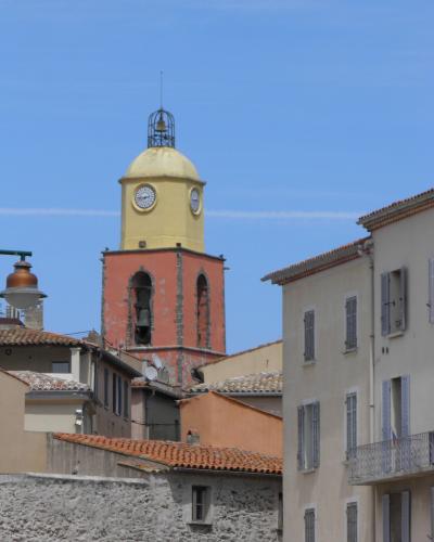 A tall, distinctive bell tower with a salmon-pink lower section and a yellow domed top featuring clocks and an open belfry, rising above terracotta-roofed buildings under a clear blue sky.