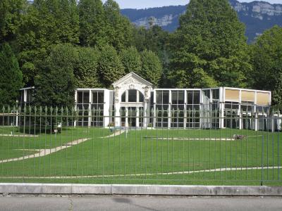 A white, classical-style pavilion with large glass windows and a central pediment, situated on a manicured lawn behind a metal fence, with forested hills and mountains in the background.