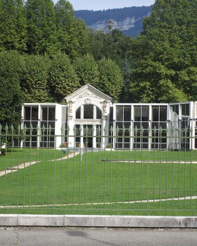 A white, classical-style pavilion with large glass windows and a central pediment, situated on a manicured lawn behind a metal fence, with forested hills and mountains in the background.
