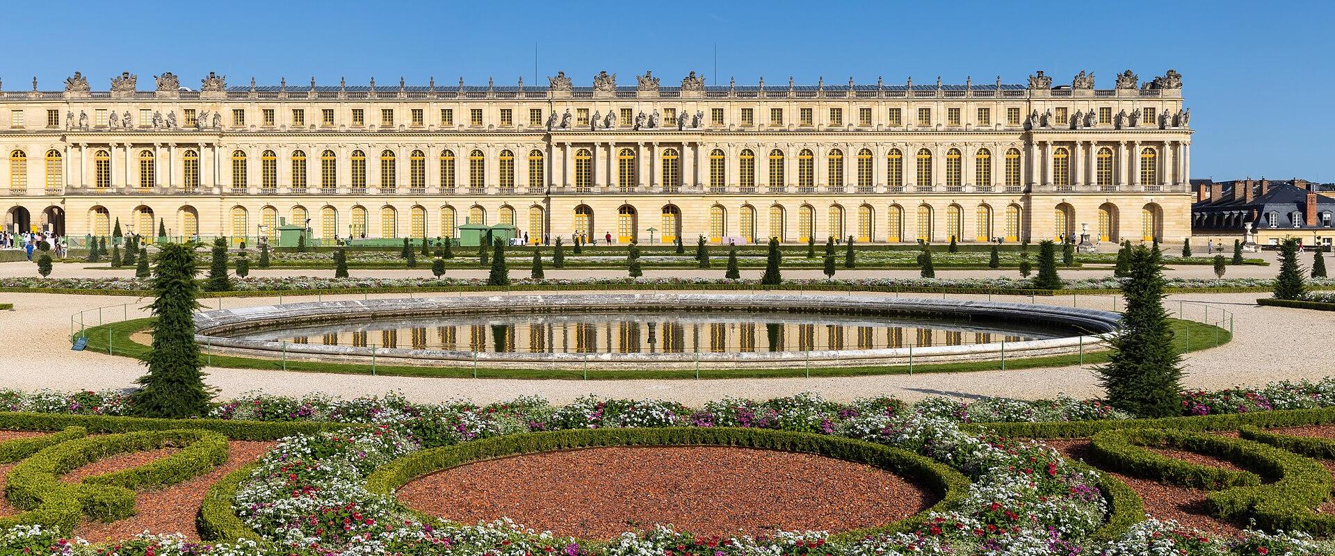 The image shows the Palace of Versailles with its formal gardens, including a circular pond and manicured flowerbeds, under a clear blue sky.