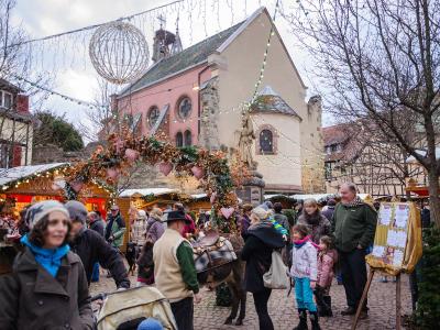 A Christmas market scene in a European town square featuring a pink church, wooden stalls, festive lights, and a crowd of people.