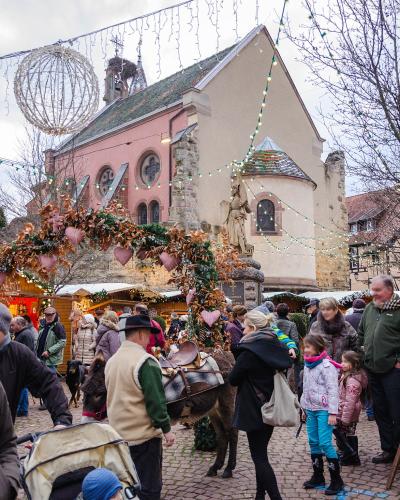 A Christmas market scene in a European town square featuring a pink church, wooden stalls, festive lights, and a crowd of people.