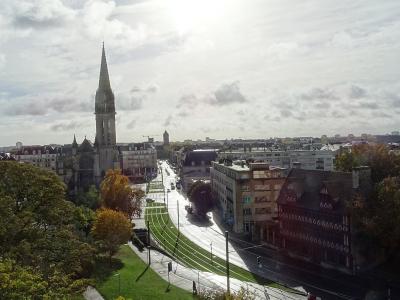A high-angle view of a French city street featuring a prominent Gothic church spire (the Église Saint-Pierre in Caen), tram tracks curving through the foreground, and surrounding urban buildings under a cloudy sky.