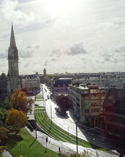 A high-angle view of a French city street featuring a prominent Gothic church spire (the Église Saint-Pierre in Caen), tram tracks curving through the foreground, and surrounding urban buildings under a cloudy sky.