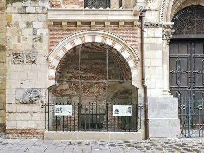 The image shows an exterior architectural detail of a historic brick and stone building, featuring a large arched opening covered by glass and a black metal fence, with informational plaques visible inside the enclosure; to the right is a heavy wo