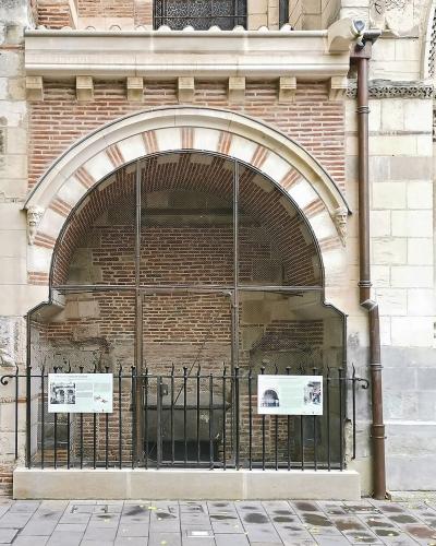 The image shows an exterior architectural detail of a historic brick and stone building, featuring a large arched opening covered by glass and a black metal fence, with informational plaques visible inside the enclosure; to the right is a heavy wo