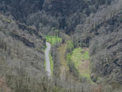 A high-angle view of a winding paved road running alongside a muddy river through a steep, wooded valley with bare trees.