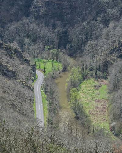 A high-angle view of a winding paved road running alongside a muddy river through a steep, wooded valley with bare trees.