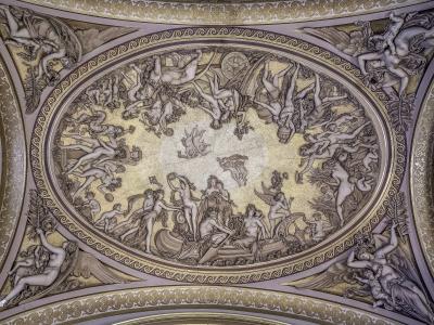 A photograph of an ornate, gilded ceiling fresco featuring a central oval medallion with mythological figures and putti, surrounded by decorative borders and corner figures.