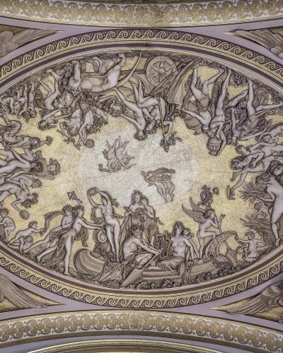 A photograph of an ornate, gilded ceiling fresco featuring a central oval medallion with mythological figures and putti, surrounded by decorative borders and corner figures.